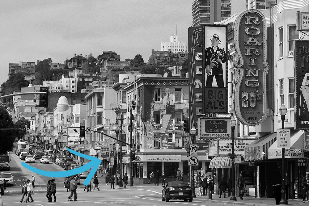 A photo looking up Broadway Street in San Francisco, with iconic neon signs in the foreground and an arrow pointing to the location of Sam's restaurant halfway up the block.