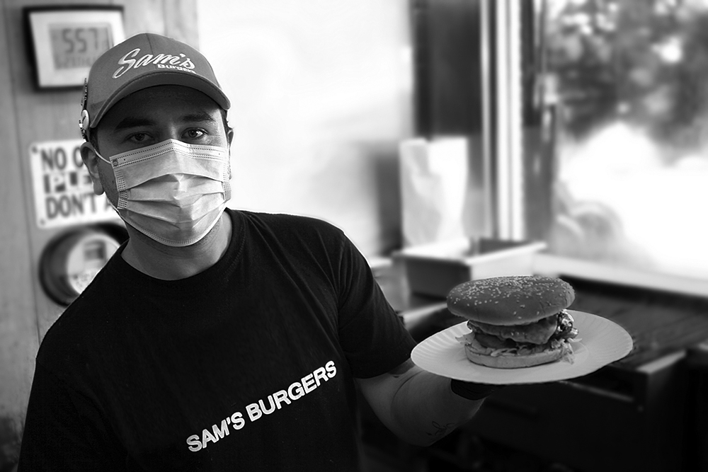 A Sam's employee wearing a face mask proudly holds up a freshly grilled hamburger.