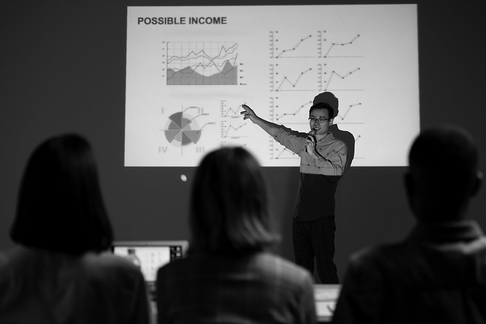 Businessman stands before a slide presentation screen pointing to a business forecast chart in a dark room with at least 3 employees.