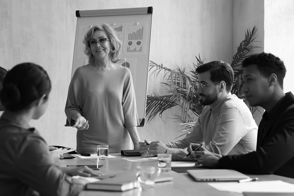 Smiling middle aged 60s businesswoman company leader standing at table, holding strategy meeting with diverse employees in boardroom.